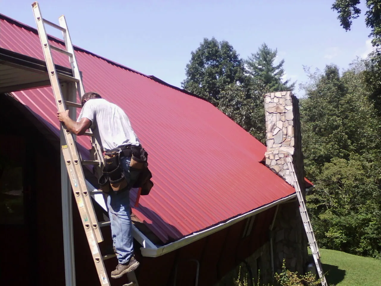 Roofer on a ladder beside a red metal roof and stone chimney — Soffit & Fascia Repair by Roofing Brothers in Atmore